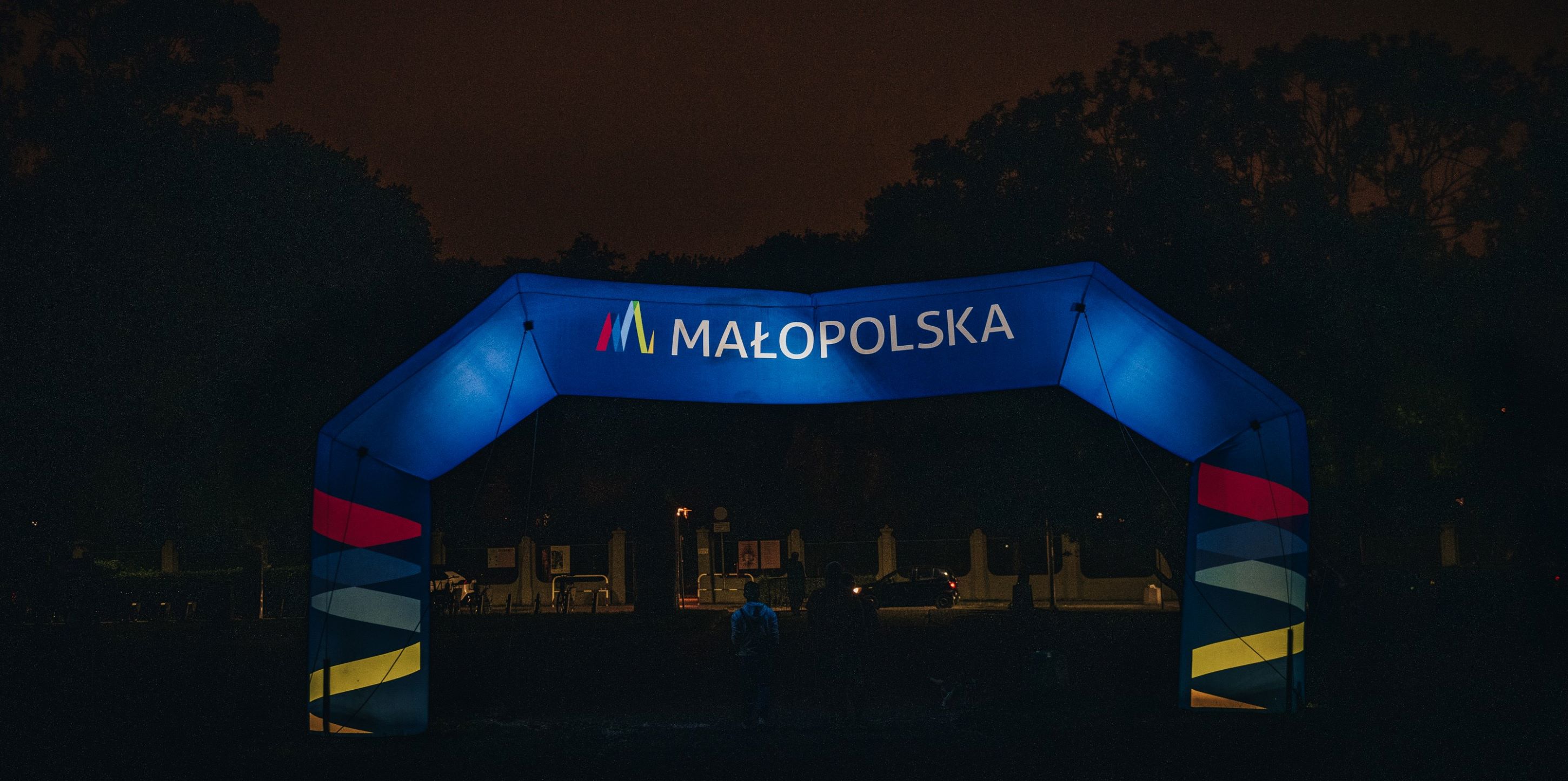 Noc, dwie osoby wychodzą przez bramę z widocznym logo małopolski.