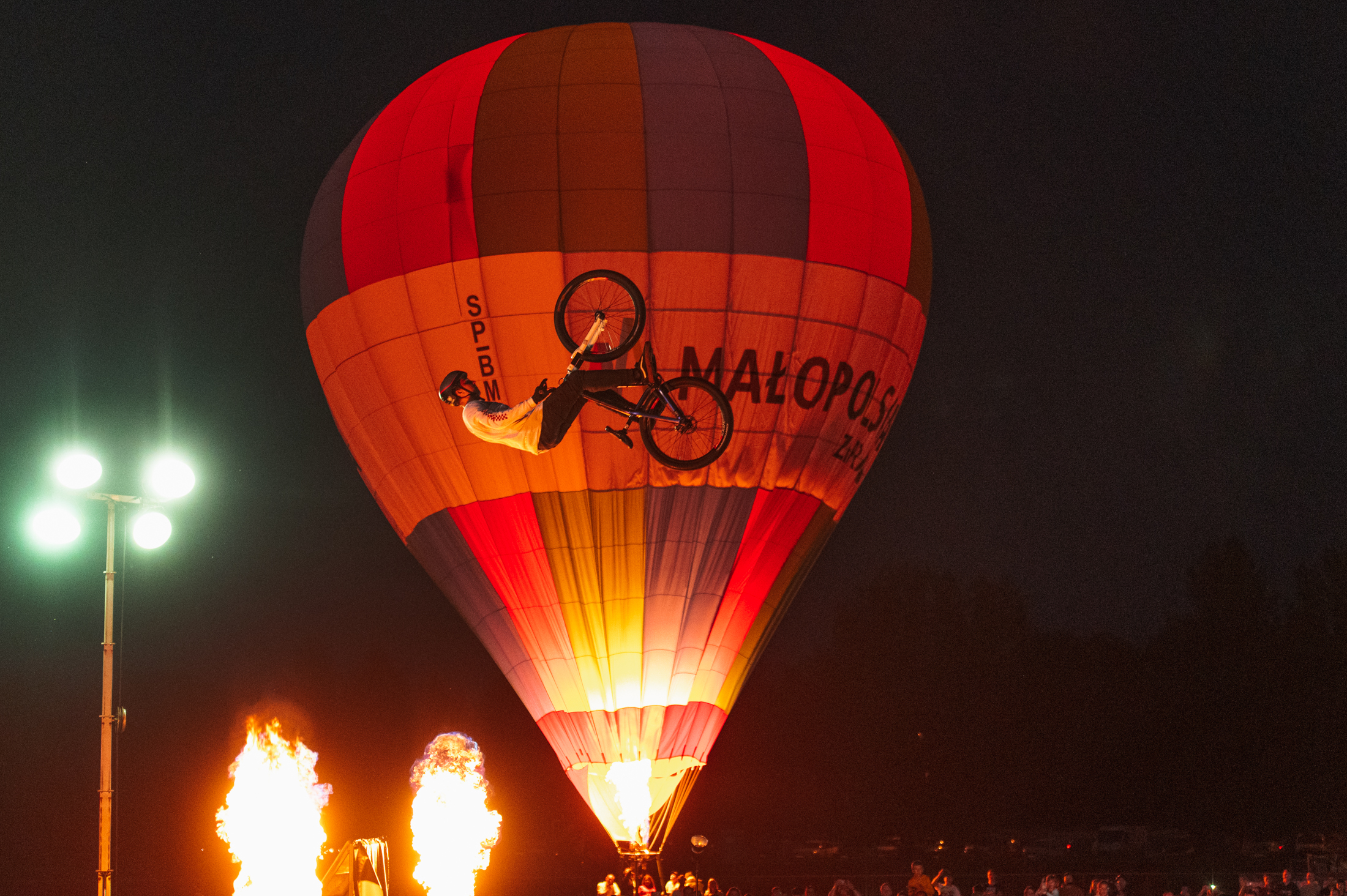 Noc oświetlona pochodniami, balon z napisem Małopolska na jego tle widać rowerzystę wykonującego salto.